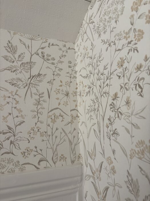 Blommig tapet i ett hörn av ett rum med sned vägg där mönsterlinjerna möts nästan perfekt, trots lutande golv och ojämna ytor.