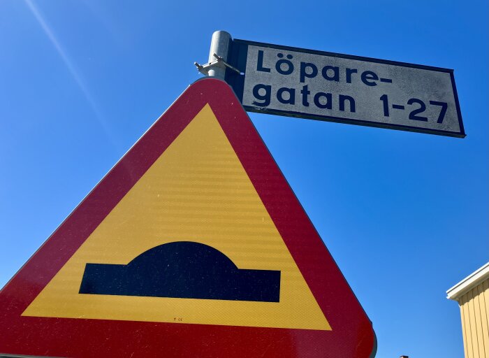 Varningsskylt för vägkudde och gattskylt med texten "Löparegatan 1-27" mot en blå himmel.