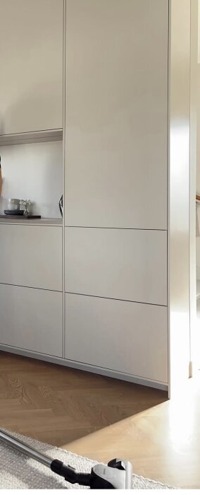 Modernt köksskåp med släta vita täckskivor som omger skåpstommar, placerat på trägolv med synlig dammsugare i förgrunden.