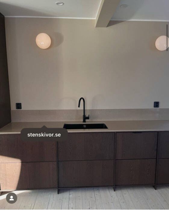 Kök med mörka träskåp, ljus köksbänk, svart blandare och diskho. Beigefärgade väggar och runda lampor på väggen.