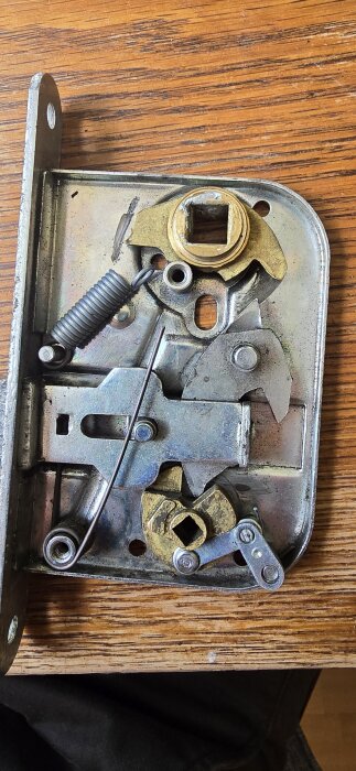 Dörrlås mekanism med synliga metalldelar och en messingsdel, placerad på ett träbord.