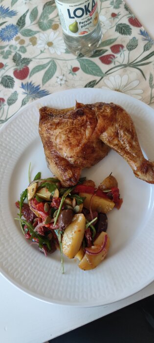 Grillad kyckling med färskpotatissallad innehållande rucola, oliver, soltorkade tomater och rödlök, serverade på en vit tallrik.