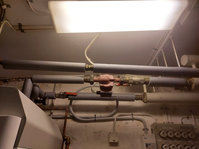 Rörsystem med isolerade rör och en shuntventil i ett pannrum under belysning.