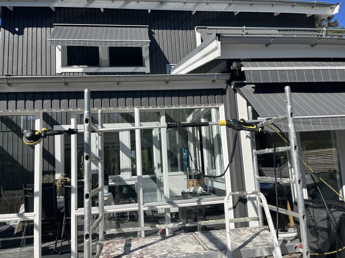 Byggställning vid glastak på uterum, rengörs med biltvättborste, synlig grå fasad och fönstermarkiser.