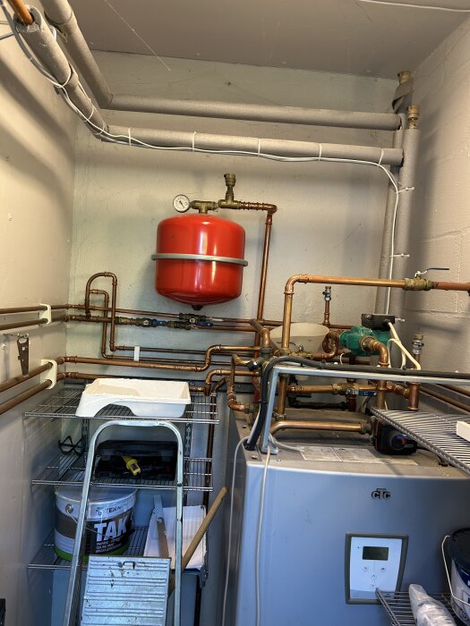 Värmesystem med kopparrör, expansionskärl och pump i ett teknikrum; stege och byggmaterial synliga.