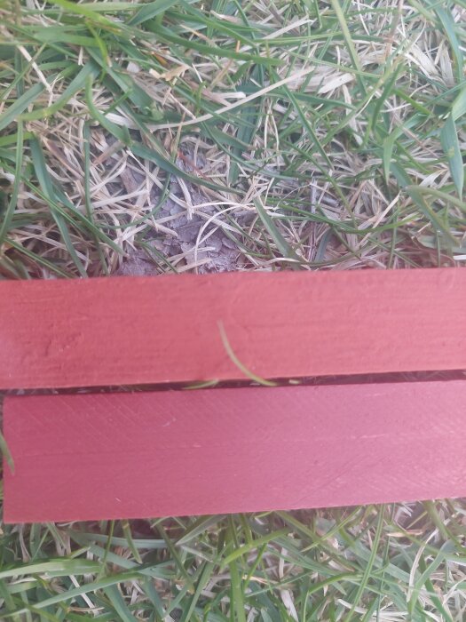 Två brädor med olika röda nyanser ligger på gräs. Den övre är äkta faluröd, den nedre 5040y80R, visar färgskillnad.