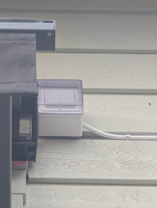 Strömbrytare installerad utomhus på trävägg, nära markis. Kapslad i plastbox, med vit kabel synlig.