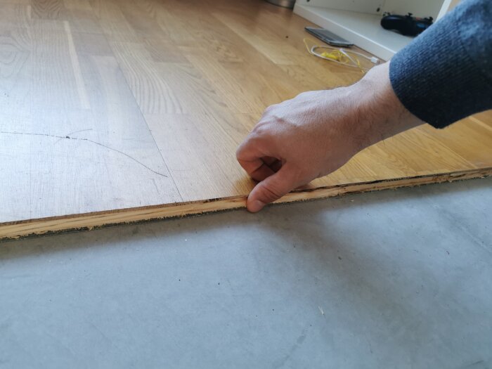 En hand lyfter en bit parkettgolv för att visa tjockleken och strukturen på brädorna ovanpå ett cementgolv.
