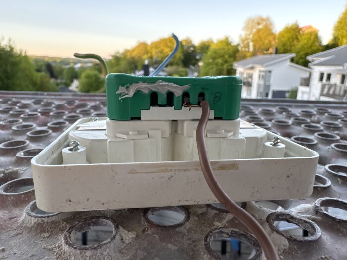 En grön kopplingsdosa med inkopplade elledningar, placerad på en perforerad yta med en vy av bostadsområde i bakgrunden.