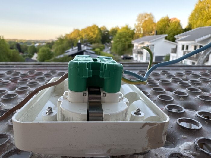 Elektrisk anslutning med gröna och blå kablar på en utomhusplats vid solnedgång, hus och träd syns i bakgrunden.