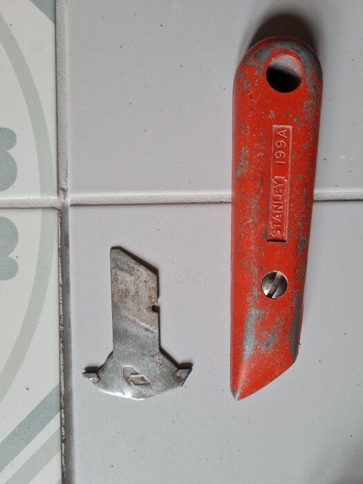 Gammalt rött verktygshandtag och metallblad på vitt kakelgolv, använd för att rensa fogar.