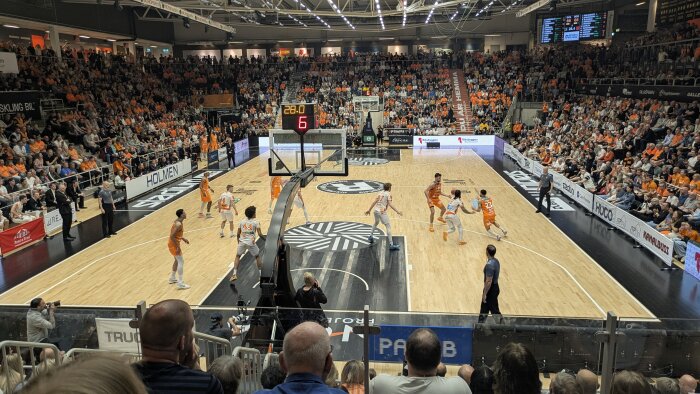 Basketmatch med fullsatta läktare, spelare i orange och vitt på plan. Resultattavlan visar slutminuter, vilket tyder på en viktig match.