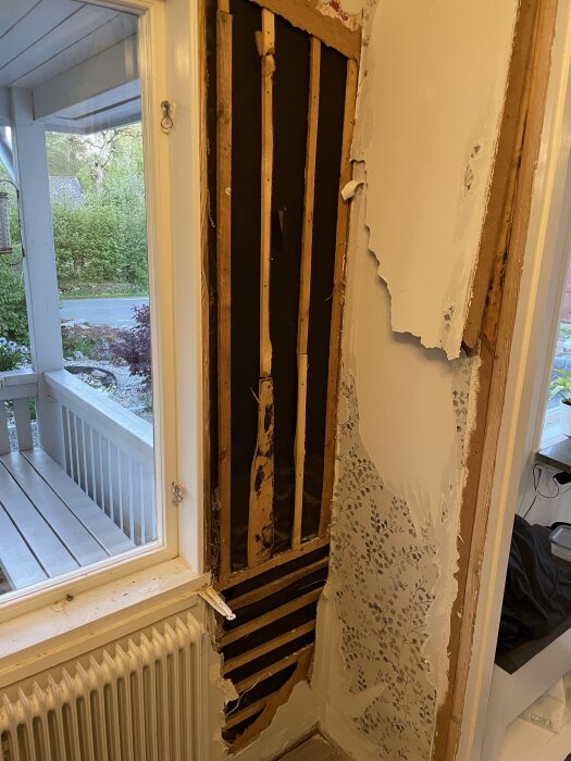 Vägg med borttagen masonit visar träreglar och svart papp vid ett fönster i en hall med trasig tapet på väggarna.