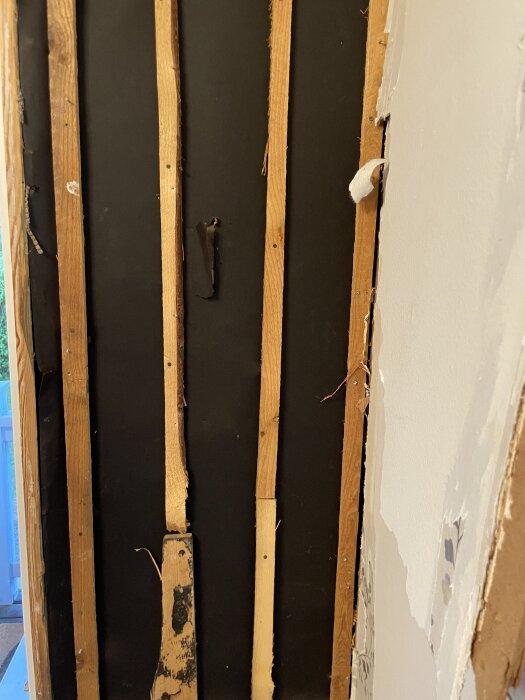 Innervägg med vertikala träreglar framför svart papp, ses delvis intakt och delvis rivet.