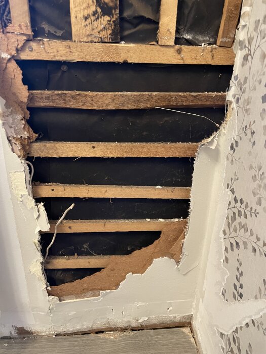 Vägg med synlig blåbetong och svart papp, delvis utriven masonit och flera lager tapet samt träribbor ovanför.