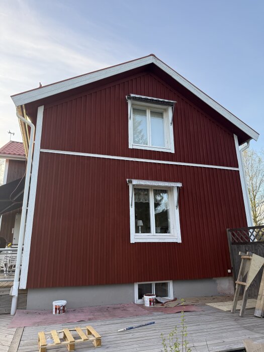 Nymålad röd husvägg med vita fönster, en vägg målad, målarburkar på trätrall.