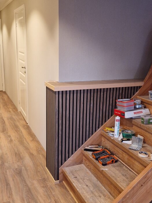 Trappa med verktyg och byggmaterial på stegen; intill trappan syns en vägg med träpaneler, del av en basturenovering.