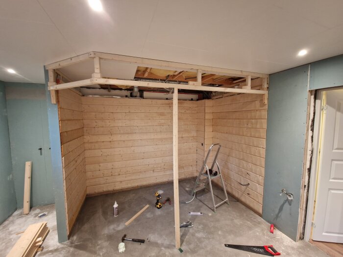 Byggarbetsplats för bastu med träpanelväggar, takkonsol under konstruktion och stege på golvet. Verktyg och träbitar liggande på betonggolv.