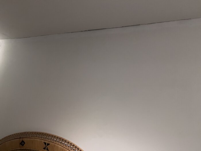Springa mellan tak och vägg i ett rum, synlig mörk linje där det inte är färdigställt, nära ett mönstrat möbelstycke.