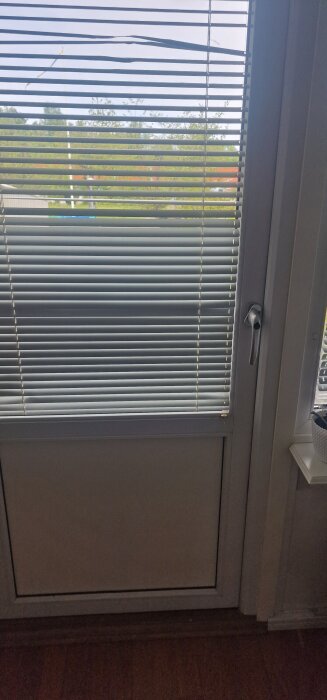Persienner med en böjd lamell framför en altandörr med utsikt över grönska och flaggstång.