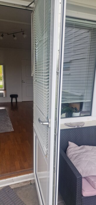 Öppen dörr med persienner, som leder från en altan till ett rum med trägolv. En korgsoffa med kudde syns bredvid dörren.