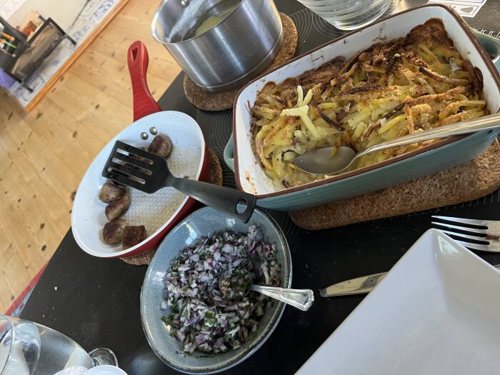 Matbord med janssons frestelse, köttbullar i stekpanna, hackad lök och gräslök i skål samt kokt potatis i kastrull.