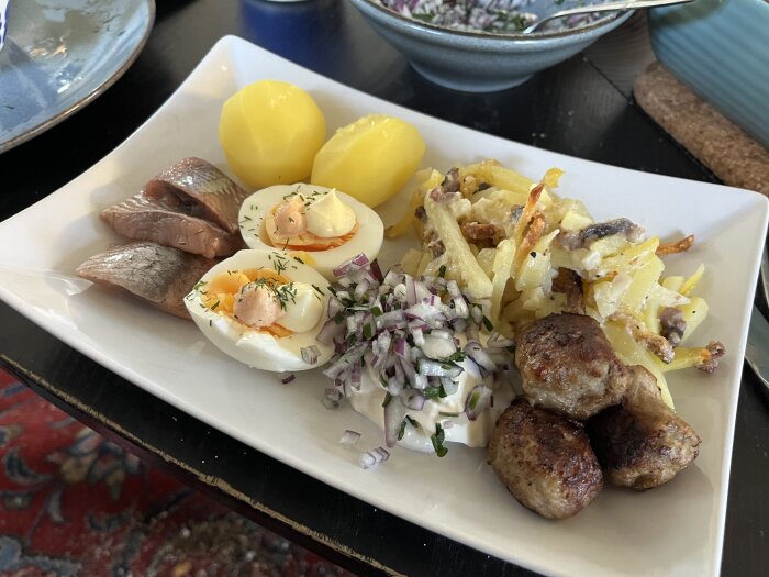 Silltallrik med matjessill, ägghalvor, janssons frestelse, köttbullar, kokt potatis, gräddfil med lök och gräslök på vitt fyrkantigt fat.