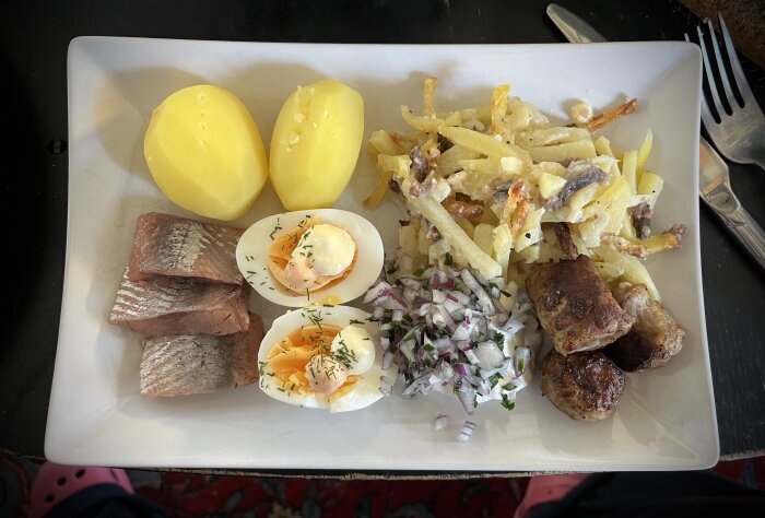 Tallrik med kokt potatis, matjessill, ägghalvor med majonnäs och dill, janssons frestelse, köttbullar, hackad lök och gräslök.