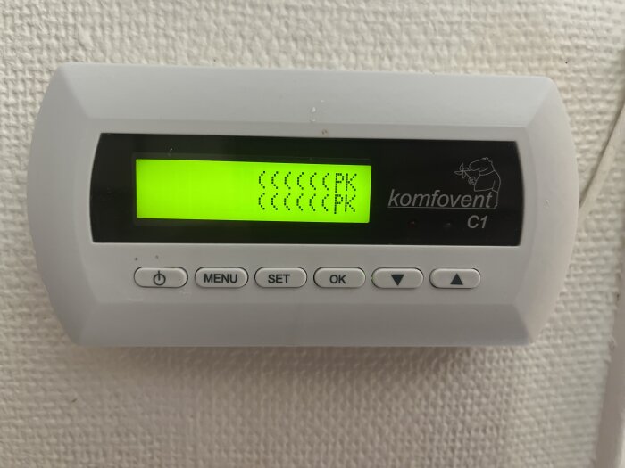 Styrpanel till Komfovent C1 med en gul display som visar symboler och text "PK". Knappar inkluderar meny, set och OK.
