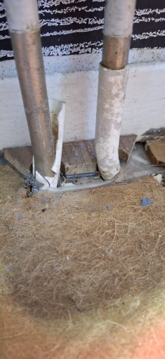 Frilagt rör i byggprojekt, med metallrör delvis täckta av vit isolering i närbild mot en vägg med träplattor vid basen.