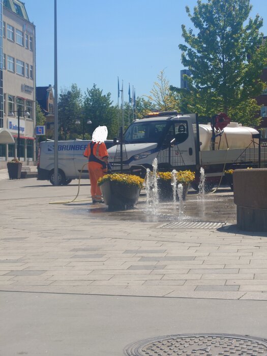 Kommunarbetare vattnar blommor i krukor bredvid en fontän, med en vattentankbil i bakgrunden på ett torg.