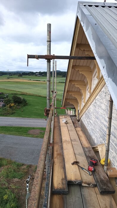 Byggställning framför ett hus med träfasad under renovering, sett från ovan. Verktyg och brädor ligger på ställningen, med utsikt över landskap i bakgrunden.