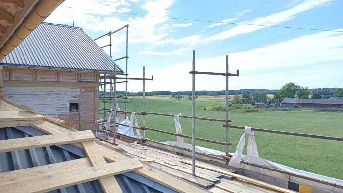 Byggnadsställning runt ett hus under renovering, med vacker utsikt över gröna fält och byggnader i bakgrunden.