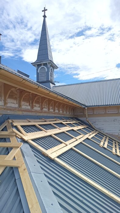 Takrenovering pågår med träställningar ovanpå ett metalltak, en kyrkspira syns i bakgrunden under en blå himmel.