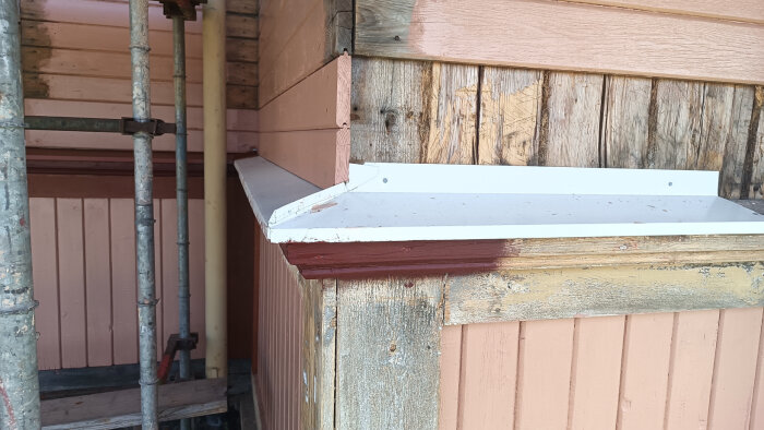 Nymålad husfasad med byggnadsställning, synligt träverk och metallkant. Vissa träpaneler är målade i olika färger, och ny plåt är installerad.