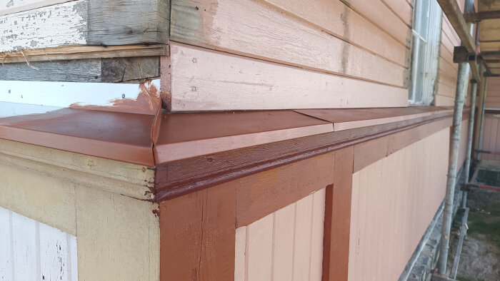 Detalj av en husfasad med ny målad panel och plåtdetaljer. Olika färglager syns, samt äldre trädetaljer som behöver underhåll.