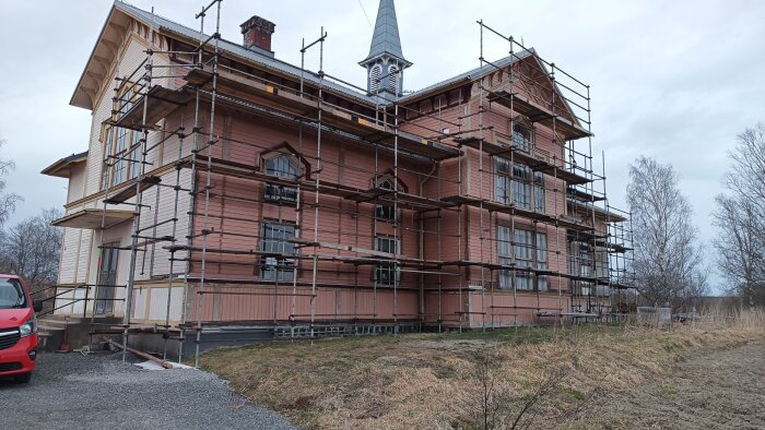 Husfasad med byggställning under renovering. Huset är delvis ommålat. Grönområde och grusplan i förgrunden, molnig himmel i bakgrunden.