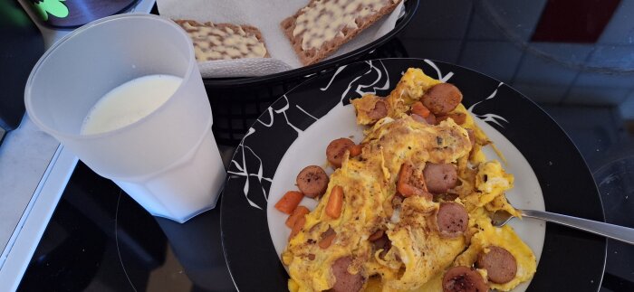 Omelett med morötter och wienerkorv på en tallrik, knäckebröd med smör och ett glas mjölk bredvid.
