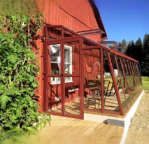 Växthus byggt med plåtprofiler, lutande glas och intill en röd träbyggnad, omgiven av grönska och utemöbler på en träveranda.