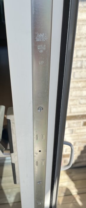 Närbild på en metalldel av en altandörr med trasig fönsterbroms, märkt med "ASSA", och en dörrhandtag som syns delvis till höger.