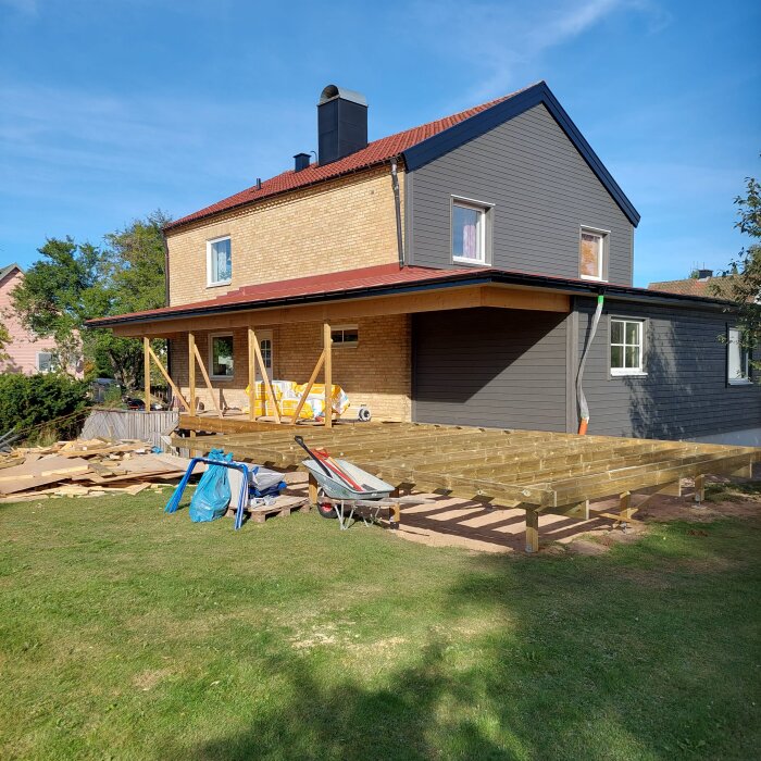 Trähus med pågående altanbygge i trädgården, slitna verktyg och material ligger runtom.