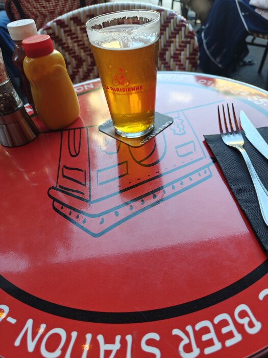 Ett glas öl på ett rött bord på en uteservering nära triumfbågen, tillsammans med senap, ketchup och bestick på en servett.
