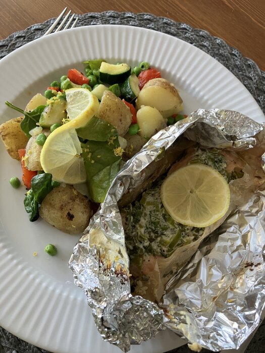 Grillad lax i folie toppad med citron bredvid färskpotatissallad med zucchini, paprika, ärtor och spenat på en tallrik.