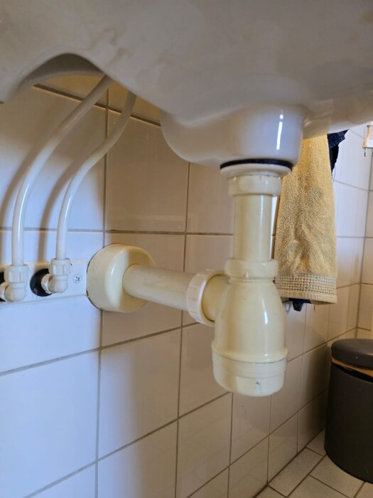 Vattenlås i plast under en vit vask i badrum, ansluten till avloppsrör i kaklat utrymme.