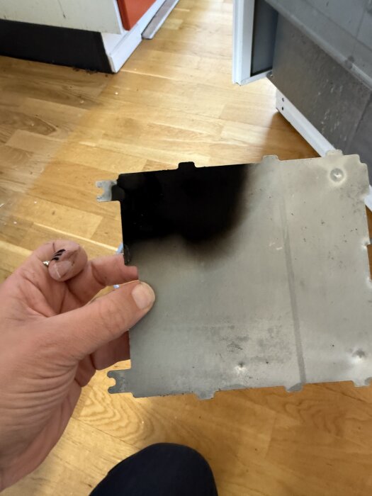 Hand som håller en metallplatta med sotfläck, tagen ur en Husqvarna spis, tecken på elbrand. Golv av trä i bakgrunden.
