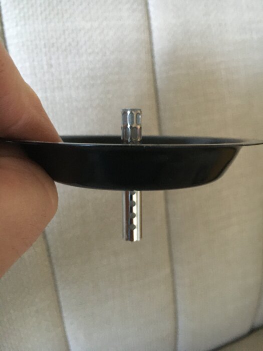 Närbild av en hand som håller en svart metallbrännare med en central cylinder och perforerad stång, mot en bakgrund av ljus tygsoffa.