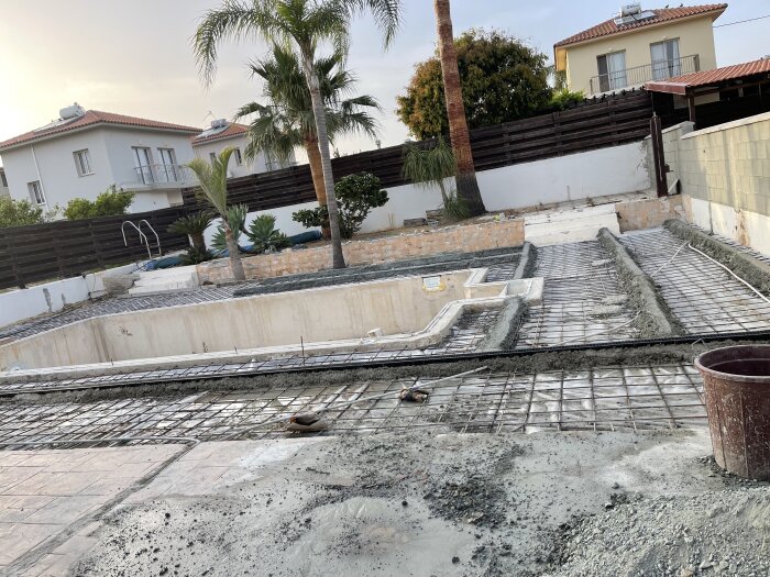 Byggarbetsplats runt en pool med betong i olika delar, armeringsjärn synliga, omgivande hus och palmer i bakgrunden.