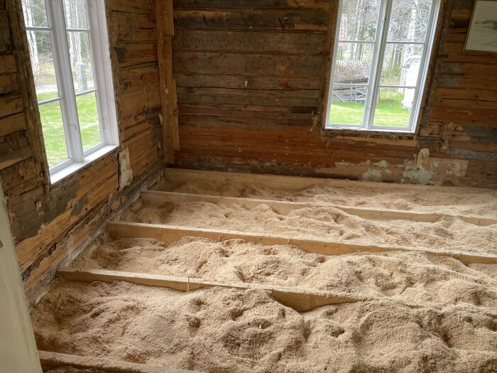 Vardagsrum under renovering med synligt trävirke och kutterspån på golvet, två stora fönster släpper in dagsljus.