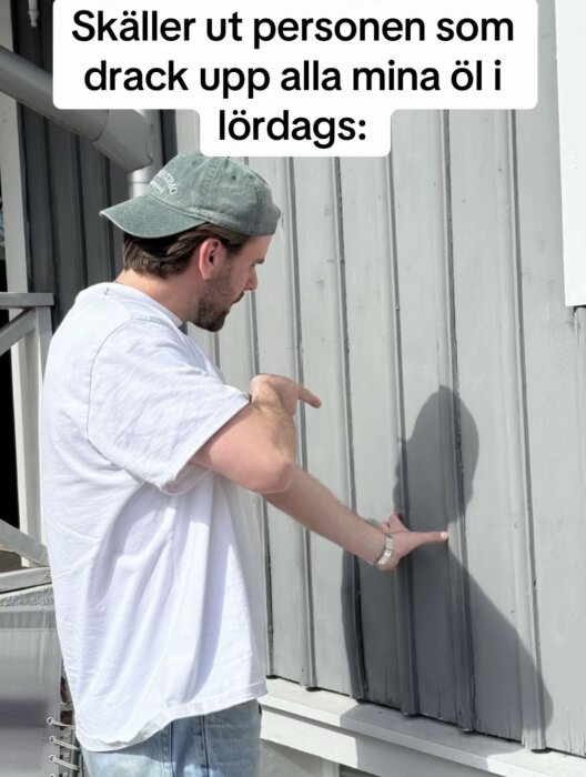 Man i vit t-shirt och keps pekar på sin egen skugga på en grå vägg, med humoristisk bildtext högst upp.