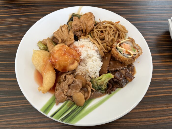 Tallrik med thaibuffémat inklusive kyckling, ris, nudlar, sushi, broccoli, och kött i sås.